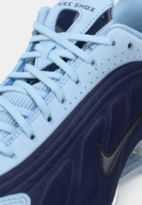 Nahaufnahme eines blauen Nike Shox Sneakers mit hellblauen Schnürsenkeln, perforiertem Obermaterial und einer dunkelblauen Sohle mit dem schwarzen Nike Swoosh-Logo.