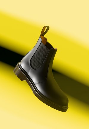 Zwarte leren Chelsea boots met getextureerde elastische zijpanelen, een gele lus en een duurzame, geprofileerde zool. De achtergrond heeft een gele gradient.