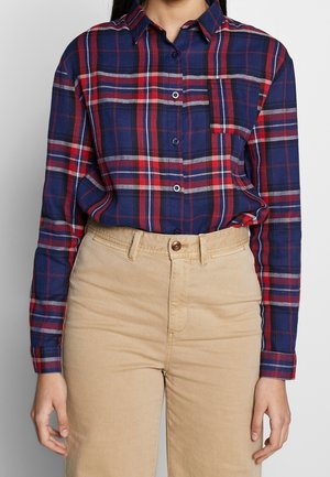 Femme portant une chemise à carreaux bleue, rouge et blanche rentrée dans un pantalon beige taille haute, debout devant un fond uni.