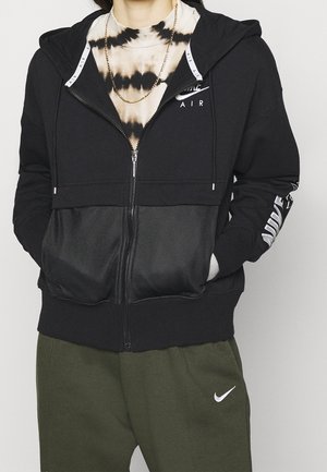 Personne portant un sweat à capuche zippé Nike Air noir par-dessus un t-shirt tie-dye noir et blanc, un pantalon Nike vert olive, et une chaîne en or autour du cou.