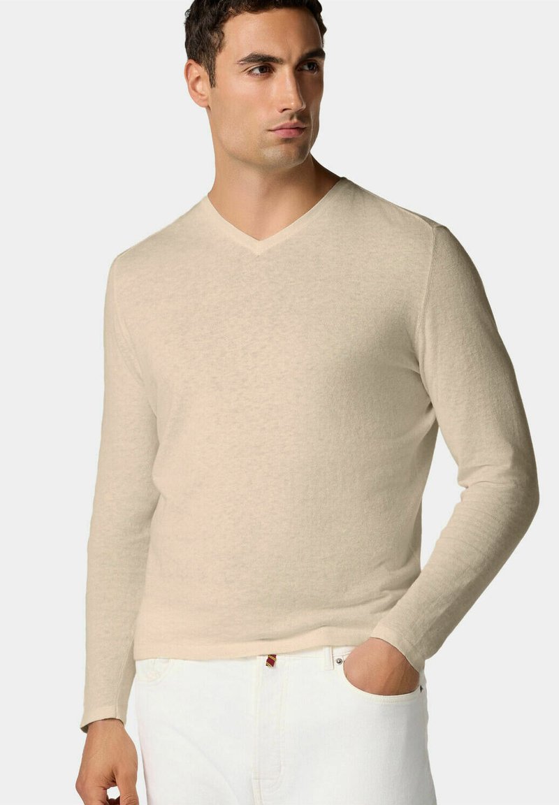 Hellbeige Langarm-V-Ausschnitt-Pullover mit weicher Textur, tailliertem Design und subtiler melierter Musterung, getragen über weißen Hosen.