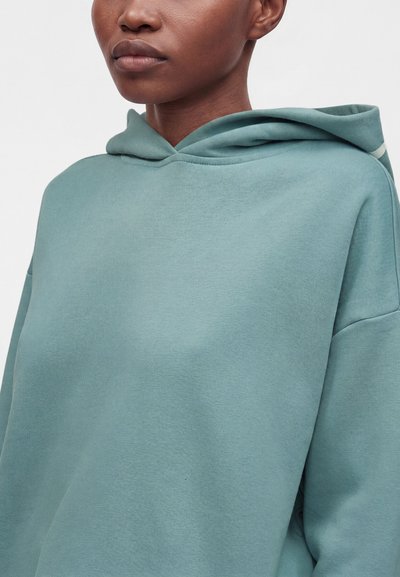 Personne portant un hoodie ample bleu sarcelle clair, vue des épaules vers le haut sur un fond uni et clair.