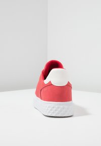 Baskets rouges avec languette de talon blanche et semelle blanche texturée, vues de dos sur une surface blanche avec un fond uni.