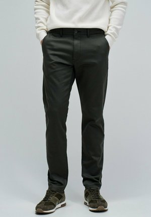 Pantalones verde oscuro de tela suave con un corte ajustado, que cuentan con bolsillos laterales y cierre de botón. Llevados con zapatillas marrones y verdes.