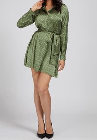 Grünes Polka-Dot-Kleid mit langen Ärmeln, Durchknopf-Front, tailliertem Gürtel und ausgestelltem Rock, aus einem glatten, glänzenden Stoff gefertigt.