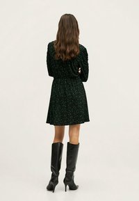 Robe noire avec un motif floral vert, taille cintrée, manches longues et ourlet évasé, associée à des bottes à talons en cuir noir montantes.