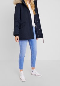 Navy blue puffer jacket with a faux fur hood, front pockets, and ribbed cuffs, worn over a white shirt and light blue jeans.