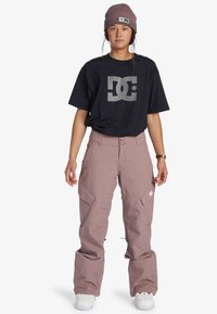 Schwarzes T-Shirt mit grauem DC-Logo, hellrosa Schneehose mit Taschen und weißen Turnschuhen. Model trägt eine braune Strickmütze.