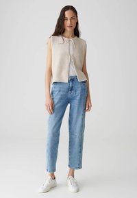 Gilet senza maniche beige in maglia con chiusura a coulisse, indossato sopra una maglietta bianca e abbinato a jeans azzurri chiari e sneakers bianche.