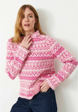 Kvinde med lyst brunt hår iført en pink og hvid mønstret sweater, poserer med den ene hånd nær sin nakke og den anden i sin jeanslomme.
