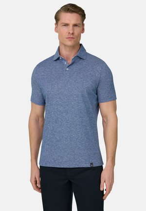 Homme portant un polo à manches courtes bleu avec des boutons et un pantalon noir, debout devant un fond clair uni.