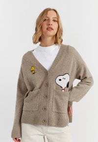Cardigan lavorato a maglia beige con scollo a V, con un design di Snoopy e Woodstock. Ha due tasche frontali e cinque bottoni marroni.