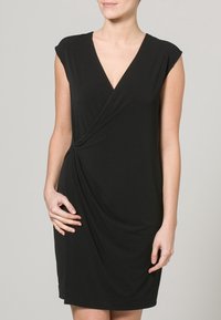 Černé šaty do půli kolen vyrobené z měkké látky, s výstřihem do V, krátkými rukávy, kroutícími se detaily v přední části a přiléhavým střihem.