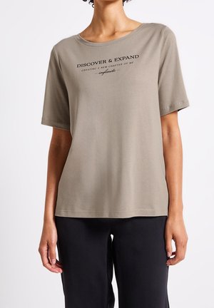 Frau trägt ein taupefarbenes kurzärmeliges T-Shirt mit dem Text „DISCOVER & EXPAND CREATING A NEW CHAPTER OF ME infinite“ und schwarze Hose.
