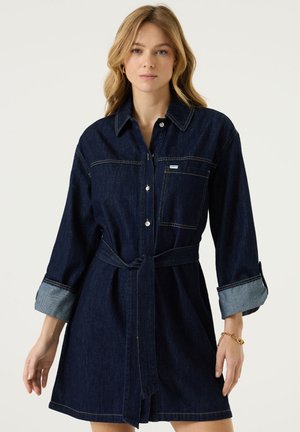 Vrouw met golvend blond haar draagt een donkerblauwe denim overhemdjurk met riem en opgerolde mouwen, staand tegen een effen lichte achtergrond.