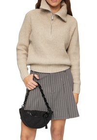 Beżowy, dziany sweter z wysokim kołnierzem i półzamkiem, w połączeniu z szarym spódnicą w paski. Trzyma małą czarną torebkę z łańcuchowym uchwytem.