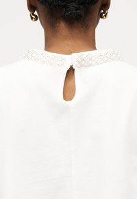 Vue de dos d'une personne aux cheveux tressés portant un haut blanc avec un col orné de perles et une découpe en forme de trou de serrure au niveau de l'encolure.