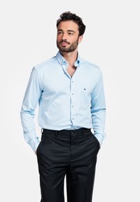 Man met donker haar en een baard, gekleed in een lichtblauw overhemd met knopen dat in zwarte broeken is gestoken, met zijn handen in zijn zakken, kijkend opzij.