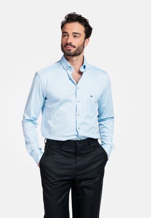 Mann mit dunklen Haaren und Bart, der ein hellblaues, in die schwarze Hose gestecktes Hemd trägt, die Hände in den Taschen, und zur Seite schaut.