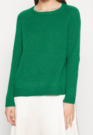 Femme portant un pull vert en maille à manches longues et une jupe blanche, montrée du cou aux hanches sur un fond uni.