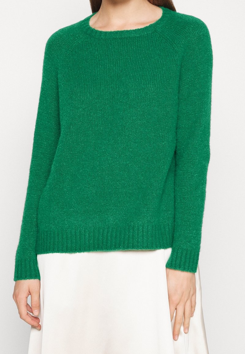 Femme portant un pull vert en maille à manches longues et une jupe blanche, montrée du cou aux hanches sur un fond uni.
