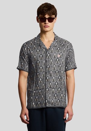 Ung mand iført mørke solbriller og en kortærmet skjorte med knapper og sort og beige blomsterprint.