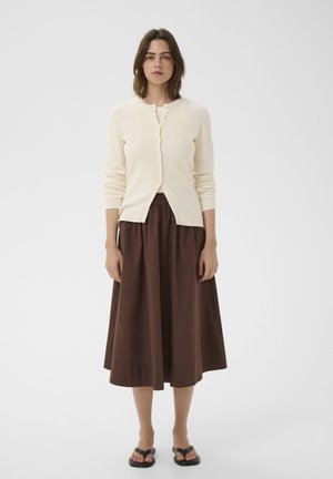 Femme debout portant un cardigan crème boutonné, une jupe midi marron et des sandales noires, sur un fond clair uni.