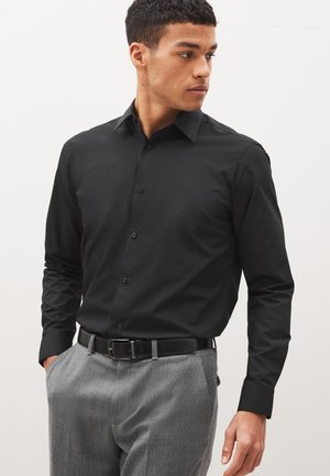 Homme portant une chemise noire à manches longues boutonnée, rentrée dans un pantalon gris avec une ceinture noire, regardant vers sa gauche.