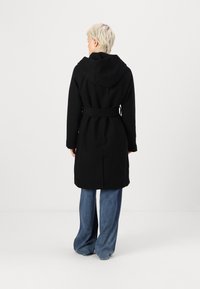 Abrigo negro con capucha, de diseño de longitud media y tejido texturizado; llevado con pantalones de mezclilla de pierna ancha.