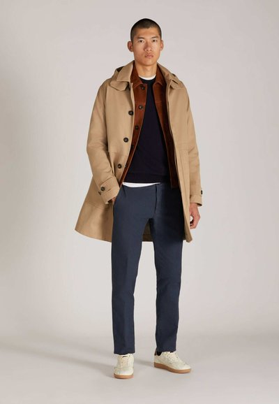 Beiger Regenmantel mit Kapuze und schwarzen Knöpfen, über einer braunen Jacke und einem marineblauen Pullover. Kombiniert mit marineblauen Hosen und weißen Sneakers.