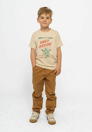 Beige t-shirt met groene en rode print met een pizzaafbeelding en tekst, gecombineerd met een paar bruine broeken en lichte sneakers met crèmekleurige accenten.