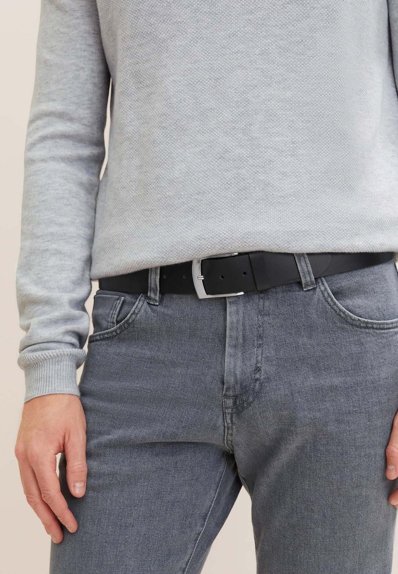 Ceinture en cuir noir avec une boucle rectangulaire en argent, portée par-dessus un jean en denim gris, associée à un pull texturé gris clair.
