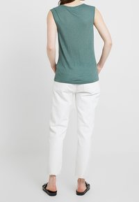 Zielona bezrękawnik o gładkiej fakturze w połączeniu z białymi dżinsami w prostym kroju. Stylizacja uzupełniona czarnymi płaskimi sandałami z ozdobnymi akcentami.