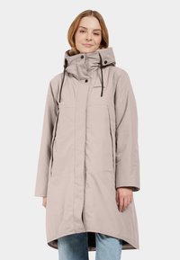 Donna che indossa un cappotto lungo beige con cappuccio e lacci neri, e jeans azzurri, in piedi contro uno sfondo bianco semplice.