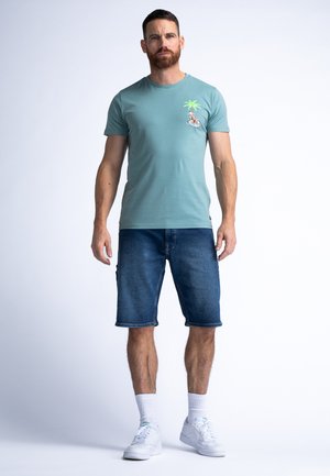 Lichtblauw t-shirt met een groene palmboom en motorfiets afbeelding. Gepaard met donkere denim shorts en witte sneakers met witte sokken.