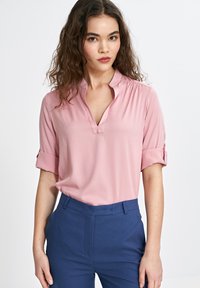 Jasnoróżowa bluzka z dekoltem w serek i krótkimi, zrolowanymi rękawami, wykonana z gładkiego materiału. W połączeniu z dopasowanymi granatowymi spodniami.