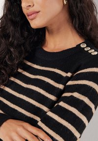 Femme aux longs cheveux bouclés portant un pull rayé noir et beige avec des boutons dorés sur l'épaule et des boucles d'oreilles anneaux torsadés dorées.