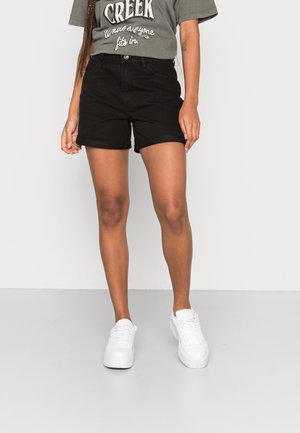 Kvinde iført sorte manchetshorts, grå t-shirt med grafik og hvide sneakers mod en ensfarvet lys baggrund.