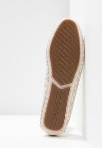 Bēšas slīdšanas apavi ar teksturētu auduma augšdaļu, gumijas zole ar zibens formas rakstu un diskrētu logo uz papēža.