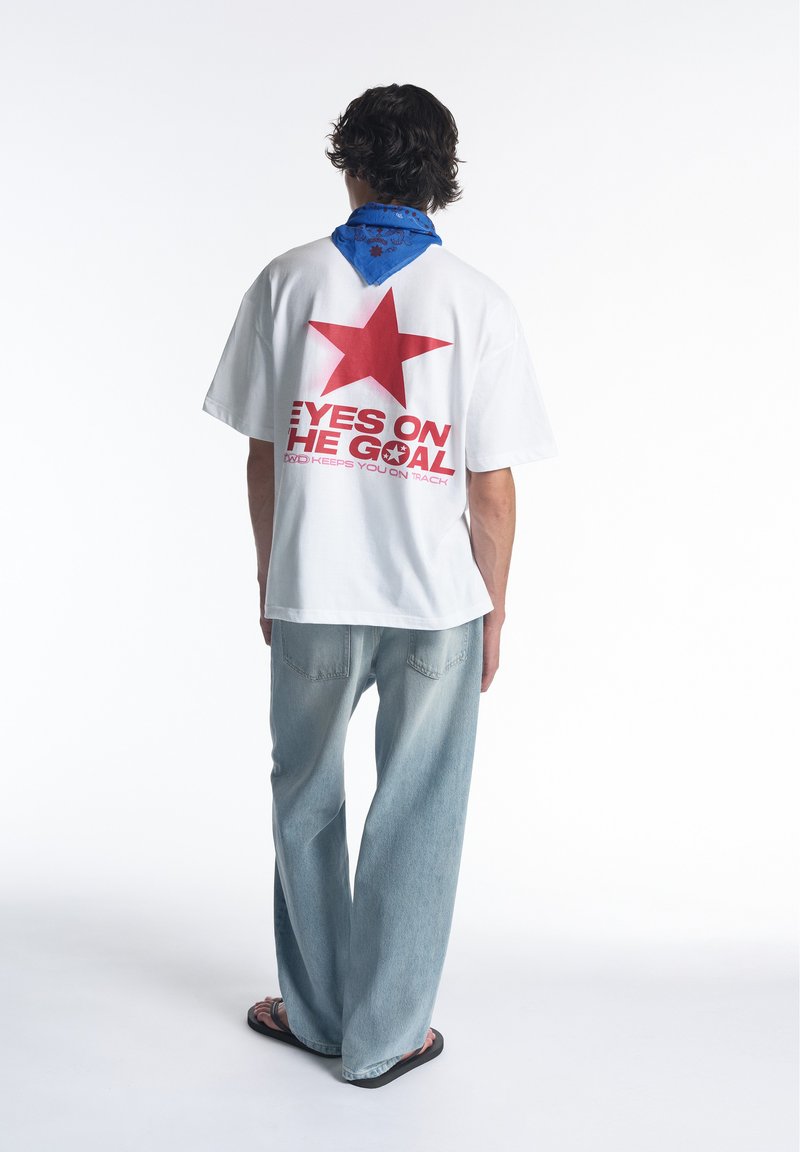 Personne portant un t-shirt blanc avec une étoile rouge et le texte "Eyes on the goal" dans le dos, bandana bleu, jean clair et sandales noires, debout dos à nous.