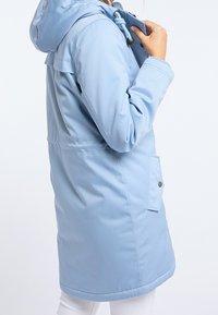 Veste bleu clair à manches longues avec capuche, dotée d'un cordon de serrage, de poches avant à boutons et d'une coupe structurée. Texture douce et finition lisse.