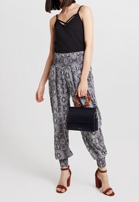 Schwarzes ärmelloses Top mit gekreuzten Trägern, gemusterte schwarz-weiße Haremshosen, dunkle Handtasche mit Holzgriff und rote Blockabsatzsandalen.