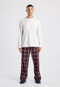 Witte shirt met lange mouwen, relaxte pasvorm. Bordeau roze geruite pyjamabroek met witte en groene accenten. Blauwe slippers. Eenvoudig, casual ontwerp.