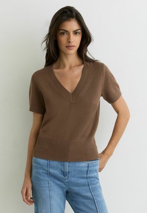 Ung kvinde med langt mørkt hår iført en brun kortærmet V-hals sweater og lyseblå jeans, stående mod en hvid væg.