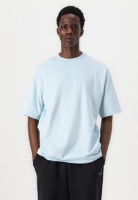 Světle modré oversized bavlněné tričko s krátkými rukávy a kulatým výstřihem. Obsahuje jemné logo na hrudi. Kombinováno s tmavými kalhotami.