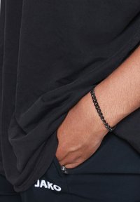 Una maglietta di cotone nera drappeggiata su pantaloni blu navy. Un bracciale a catena nera adorna una mano che riposa in tasca, mostrando una superficie liscia.