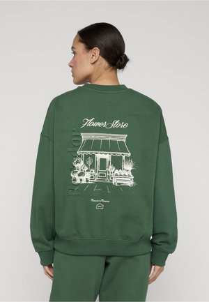 Persona con felpa verde con un disegno a linee bianche di un negozio di fiori, piante e il testo "Flower Store" sul retro.