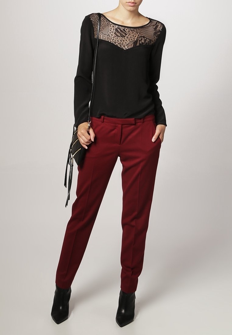 Femme portant un haut noir à manches longues avec un détail en dentelle, un pantalon bordeaux sur mesure, des bottines noires et un sac à épaule noir.