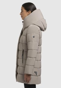 Manteau doudoune beige clair avec un col montant, design matelassé, poche latérale zippée et écusson logo sur la manche. Texture de tissu lisse.