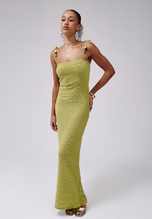 Femme portant une robe texturée vert citron sans manches avec des bretelles à nouer et des talons ouverts dorés, debout sur un fond clair uni.